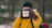 A person dressed in a bright yellow jacket holds a white mug with the words 'Adventure Begins' written on it. The background is blurred, suggesting a natural, outdoor setting, possibly a forest. The person is wearing brown gloves and a black beanie.