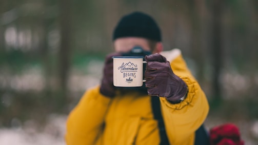 A person dressed in a bright yellow jacket holds a white mug with the words 'Adventure Begins' written on it. The background is blurred, suggesting a natural, outdoor setting, possibly a forest. The person is wearing brown gloves and a black beanie.