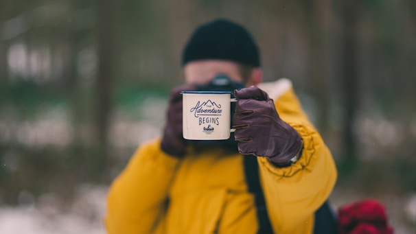 A person dressed in a bright yellow jacket holds a white mug with the words 'Adventure Begins' written on it. The background is blurred, suggesting a natural, outdoor setting, possibly a forest. The person is wearing brown gloves and a black beanie.