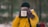 A person dressed in a bright yellow jacket holds a white mug with the words 'Adventure Begins' written on it. The background is blurred, suggesting a natural, outdoor setting, possibly a forest. The person is wearing brown gloves and a black beanie.