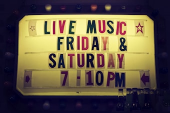 A sign with a yellow background features the text 'LIVE MUSIC' in large letters, followed by 'FRIDAY & SATURDAY 7-10PM' in a combination of red and green letters. Decorative star icons are on each side, accompanied by directional arrows. The sign is bordered by multi-colored bulbs, creating a vintage look. A few beverage bottles are visible at the bottom of the image.