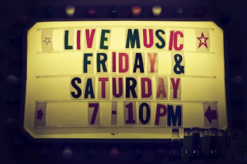 A sign with a yellow background features the text 'LIVE MUSIC' in large letters, followed by 'FRIDAY & SATURDAY 7-10PM' in a combination of red and green letters. Decorative star icons are on each side, accompanied by directional arrows. The sign is bordered by multi-colored bulbs, creating a vintage look. A few beverage bottles are visible at the bottom of the image.