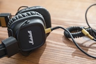 A pair of black over-ear headphones with a coiled cable and gold connectors rests on a wooden surface. The headphones have a textured design with a prominent logo on the ear cups.