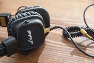 A pair of black over-ear headphones with a coiled cable and gold connectors rests on a wooden surface. The headphones have a textured design with a prominent logo on the ear cups.
