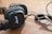 A pair of black over-ear headphones with a coiled cable and gold connectors rests on a wooden surface. The headphones have a textured design with a prominent logo on the ear cups.