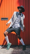 A fashionable person poses in front of a bright orange backdrop. They are wearing a grey jacket over a white shirt with a bowtie, black shorts, and green sneakers. Accessories include a black hat and glasses. The combination of clothing gives a modern yet classic vibe.