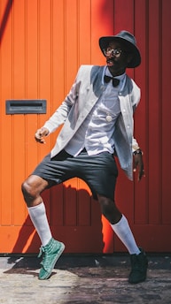 A fashionable person poses in front of a bright orange backdrop. They are wearing a grey jacket over a white shirt with a bowtie, black shorts, and green sneakers. Accessories include a black hat and glasses. The combination of clothing gives a modern yet classic vibe.