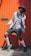 A fashionable person poses in front of a bright orange backdrop. They are wearing a grey jacket over a white shirt with a bowtie, black shorts, and green sneakers. Accessories include a black hat and glasses. The combination of clothing gives a modern yet classic vibe.