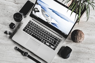 A workspace setup with a silver laptop displaying a scenic wallpaper, surrounded by various items including a smartphone, camera lens, wristwatch, wireless earbuds, and a computer mouse. A small plant and a coconut add natural elements to the minimalist white surface.