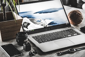 A workspace featuring an open laptop displaying a serene coastal scene with a modern building. Surrounding the laptop are various items including a smartphone, a camera lens, a small camera, a watch, a pair of wireless earbuds, a potted plant, and a coconut.