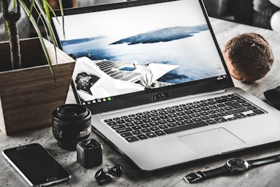 A workspace featuring an open laptop displaying a serene coastal scene with a modern building. Surrounding the laptop are various items including a smartphone, a camera lens, a small camera, a watch, a pair of wireless earbuds, a potted plant, and a coconut.