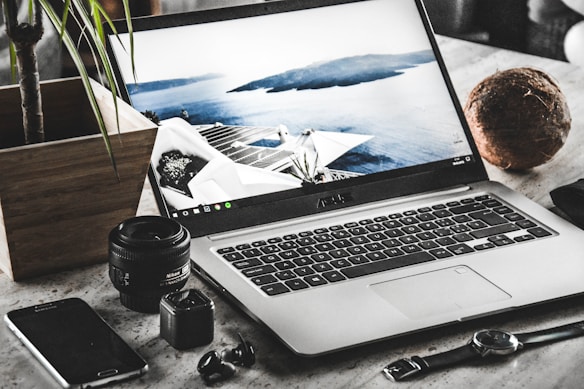 A workspace featuring an open laptop displaying a serene coastal scene with a modern building. Surrounding the laptop are various items including a smartphone, a camera lens, a small camera, a watch, a pair of wireless earbuds, a potted plant, and a coconut.