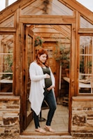 A pregnant woman stands in the doorway of a wooden greenhouse, gently holding her belly with one hand while the other hand rests on her side. She is wearing a green top, blue jeans, a white cardigan, and tan sandals. Inside the greenhouse, there are plants in pots and wooden furniture.