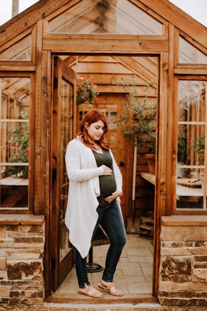 A pregnant woman stands in the doorway of a wooden greenhouse, gently holding her belly with one hand while the other hand rests on her side. She is wearing a green top, blue jeans, a white cardigan, and tan sandals. Inside the greenhouse, there are plants in pots and wooden furniture.