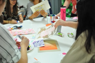 A group of people are sitting around a table engaged in an art or craft activity. Various art supplies, including colored pens, paper, and glue sticks, are scattered on the table. Participants are focused on creating and discussing their projects, and vibrant patterns and designs are visible.