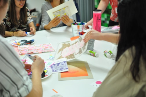 A group of people are sitting around a table engaged in an art or craft activity. Various art supplies, including colored pens, paper, and glue sticks, are scattered on the table. Participants are focused on creating and discussing their projects, and vibrant patterns and designs are visible.