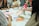 A group of people are sitting around a table engaged in an art or craft activity. Various art supplies, including colored pens, paper, and glue sticks, are scattered on the table. Participants are focused on creating and discussing their projects, and vibrant patterns and designs are visible.