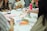 A group of people are sitting around a table engaged in an art or craft activity. Various art supplies, including colored pens, paper, and glue sticks, are scattered on the table. Participants are focused on creating and discussing their projects, and vibrant patterns and designs are visible.