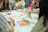 A group of people are sitting around a table engaged in an art or craft activity. Various art supplies, including colored pens, paper, and glue sticks, are scattered on the table. Participants are focused on creating and discussing their projects, and vibrant patterns and designs are visible.