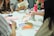A group of people are sitting around a table engaged in an art or craft activity. Various art supplies, including colored pens, paper, and glue sticks, are scattered on the table. Participants are focused on creating and discussing their projects, and vibrant patterns and designs are visible.