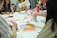 A group of people are sitting around a table engaged in an art or craft activity. Various art supplies, including colored pens, paper, and glue sticks, are scattered on the table. Participants are focused on creating and discussing their projects, and vibrant patterns and designs are visible.