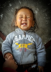 A young child with closed eyes is smiling widely, wearing a gray hoodie with the word 'CHAMPION' and some letters and designs in yellow and red. The child appears to be held by an adult whose hand is visible on the left side of the image, and they are set against a textured background.