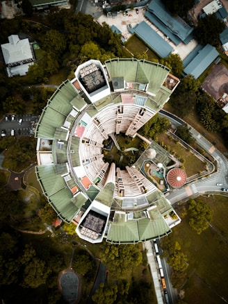 An aerial view of a circular building with a futuristic design, featuring a large central void surrounded by sections with green roofs. The structure is set amid lush greenery with roads curving around it. Nearby, there are smaller buildings and a swimming pool, with vehicles visible on the road.