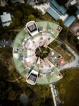 An aerial view of a circular building with a futuristic design, featuring a large central void surrounded by sections with green roofs. The structure is set amid lush greenery with roads curving around it. Nearby, there are smaller buildings and a swimming pool, with vehicles visible on the road.