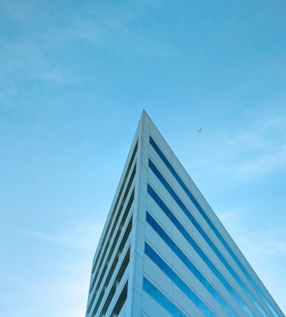 The image features a modern, triangular-shaped building with a sleek design against a clear blue sky. A small airplane is visible flying above the building, adding a sense of scale and movement.