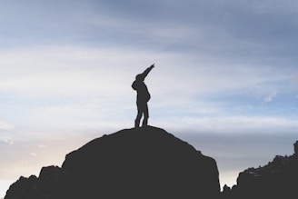 A silhouette of a person standing on top of a large rock with one arm extended upwards against a backdrop of a cloudy sky. The scene suggests a moment of achievement or victory.
