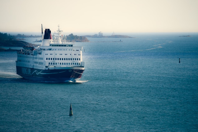 A large ferry with multiple decks navigates through calm blue waters. The ship is heading towards a cluster of small, rocky islands visible in the background. The water is slightly rippled, and the sky is hazy, suggesting a tranquil atmosphere.