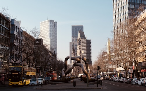 A busy urban street with modern and historical architecture. In the center, a sculptural installation stands out, surrounded by tall buildings. Bare trees line the street, and a yellow double-decker bus is among the vehicles present. The scene conveys a sense of city life and architecture.