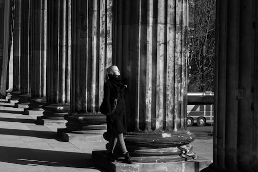 A person stands near large, classical stone columns, basking in sunlight. The scene has a striking play of light and shadow, with strong vertical lines created by the columns. The person appears calm and contemplative, wearing a dark coat and standing on a plinth. In the background, a vehicle is partially visible, framed between the columns.