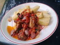 A plate of food with fried cassava pieces and a vibrant tomato-based sauce containing onions and herbs. Pieces of grilled or roasted meat are garnished atop the sauce, adding texture and flavor to the dish.