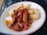 A plate of food with fried cassava pieces and a vibrant tomato-based sauce containing onions and herbs. Pieces of grilled or roasted meat are garnished atop the sauce, adding texture and flavor to the dish.