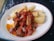 A plate of food with fried cassava pieces and a vibrant tomato-based sauce containing onions and herbs. Pieces of grilled or roasted meat are garnished atop the sauce, adding texture and flavor to the dish.