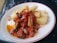 A plate of food with fried cassava pieces and a vibrant tomato-based sauce containing onions and herbs. Pieces of grilled or roasted meat are garnished atop the sauce, adding texture and flavor to the dish.