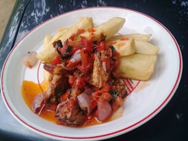 A plate of food with fried cassava pieces and a vibrant tomato-based sauce containing onions and herbs. Pieces of grilled or roasted meat are garnished atop the sauce, adding texture and flavor to the dish.