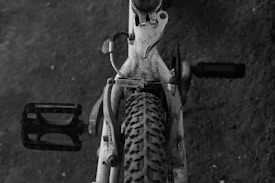 A black and white image features a close-up view of a bicycle from the top, focusing on the pedal, tire, and part of the frame. The image has a textured appearance with visible dirt on the bike.