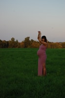 A pregnant woman stands gracefully in a lush green field, wearing a form-fitting pink dress. Her arms are raised above her head, and she is surrounded by a serene landscape with a backdrop of trees and a clear sky.
