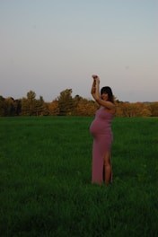 A pregnant woman stands gracefully in a lush green field, wearing a form-fitting pink dress. Her arms are raised above her head, and she is surrounded by a serene landscape with a backdrop of trees and a clear sky.