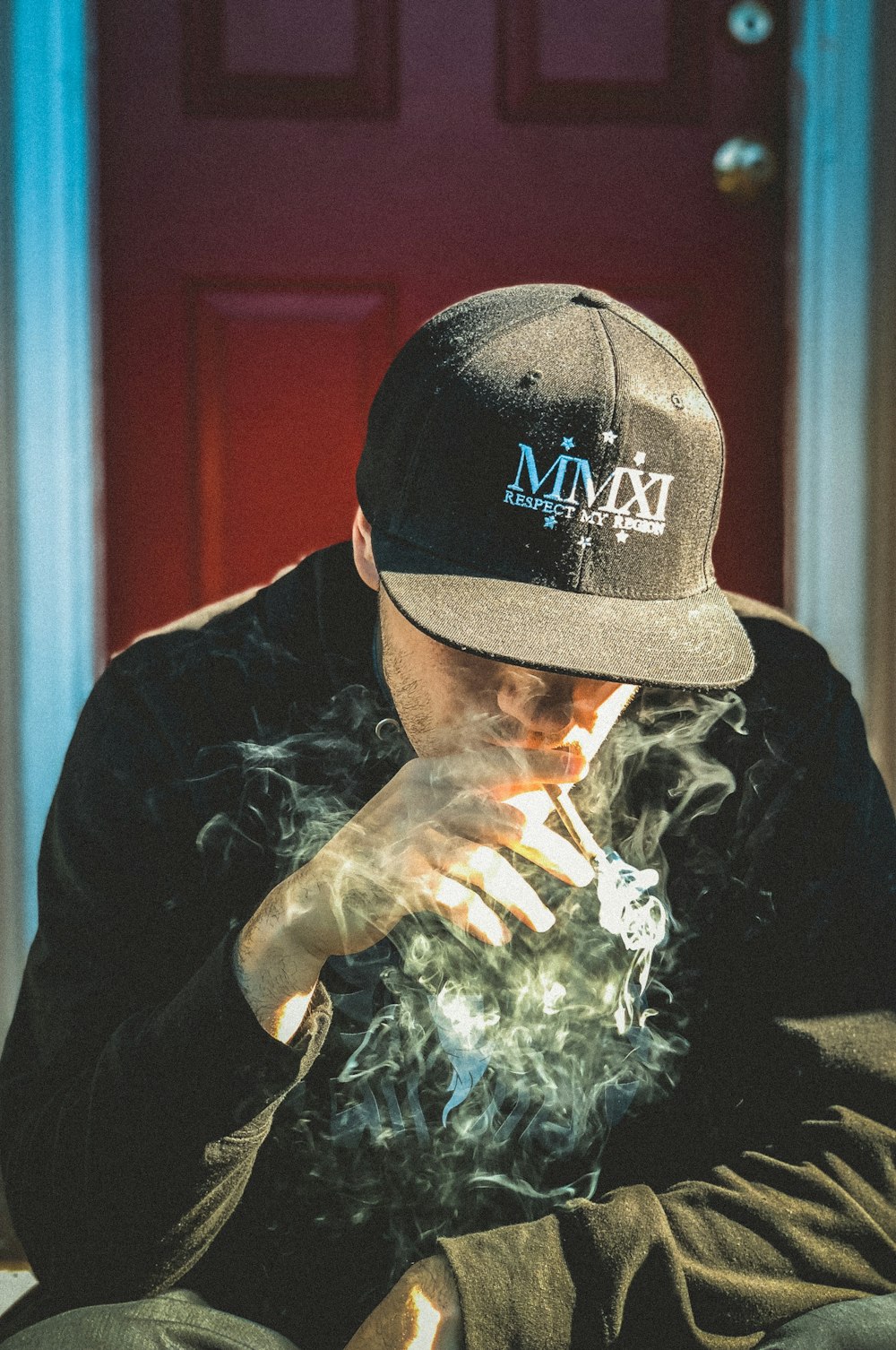 검은 모자와 셔츠를 입은 남자가 담배를 피우며 문 근처에 앉아 있습니다.