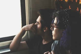Two individuals are seated next to each other, gazing out of a window. Soft lighting suggests a reflective or contemplative mood, with one person resting their chin on their hand. The focus is on their profiles and expressions, indicating a sense of calm or introspection.