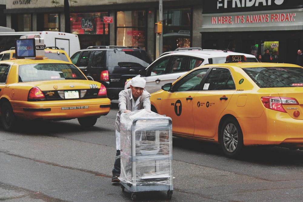 man pushing gray bus cart on road near two yellow taxi cabs