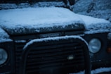 A Land Rover Defender covered in a layer of snow. The hood and front bumper are visibly blanketed with snow, highlighting the car’s rugged design. The vehicle's logo and headlights are partially covered, indicating a cold and wintry environment.