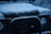 A Land Rover Defender covered in a layer of snow. The hood and front bumper are visibly blanketed with snow, highlighting the car’s rugged design. The vehicle's logo and headlights are partially covered, indicating a cold and wintry environment.
