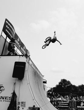 A cyclist is performing a high-flying trick on a ramp with a BMX bike, extending their arms and legs mid-air. The image captures the dynamic motion and balance against a cloudy sky. A small crowd is gathered near the ramp, some looking up to watch the thrilling action.