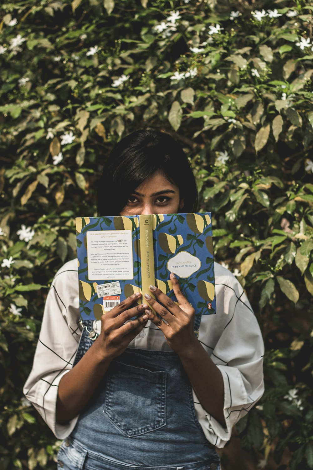 Frau, die ihr Gesicht mit einem Buch bedeckt, steht vor grünen Pflanzen