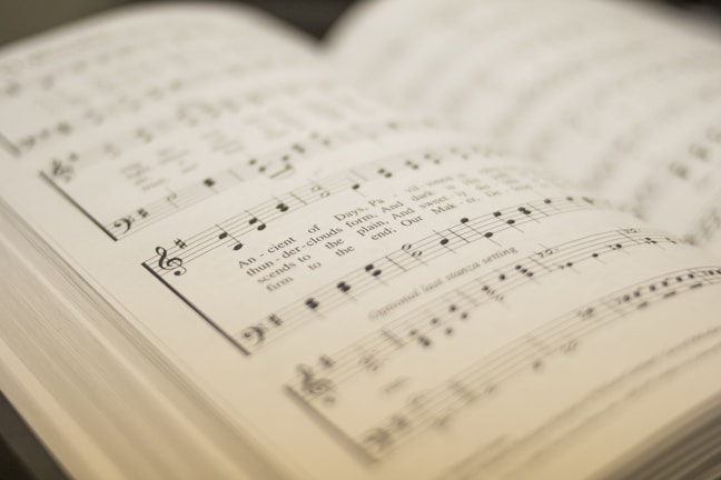 An open sheet music book displaying musical notations and lyrics. The pages are slightly blurred in the background, focusing on a section of the music with clearly visible notes and treble clef.