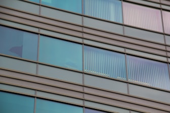 Glass windows of a modern building with horizontal metal frames and reflective surfaces. Some windows show vertical blinds partially drawn, allowing a view of interior silhouettes.